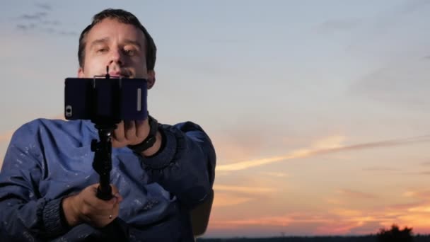 Ein junger Mann sitzt bei Sonnenuntergang und macht ein Selfie mit seinem Handy. schöner Himmel dahinter. — Stockvideo