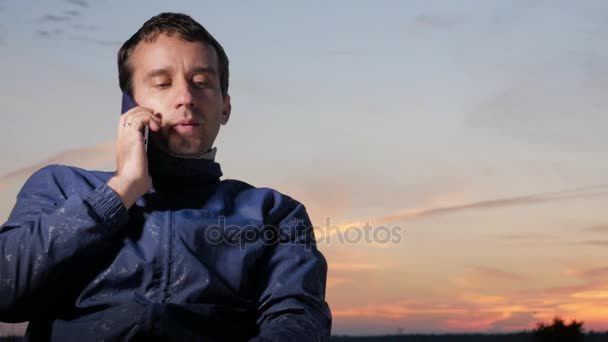 Un joven se sienta al atardecer y habla por teléfono móvil. Hermoso cielo detrás . — Vídeos de Stock