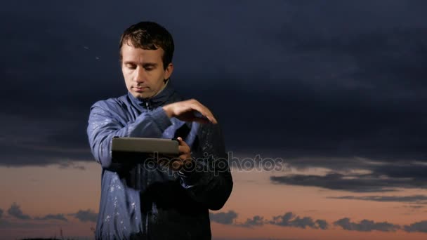 Söt ung man arbetar på en handdator i solnedgången på fältet. Vacker himmel på en bakgrund. — Stockvideo