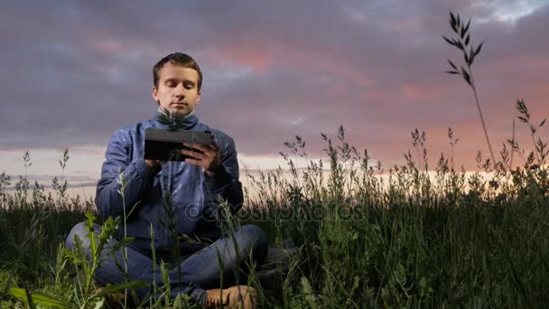 Roztomilý mladý muž pracuje na tabletovém počítači při západu slunce na hřišti. Krásná obloha na pozadí. — Stock video