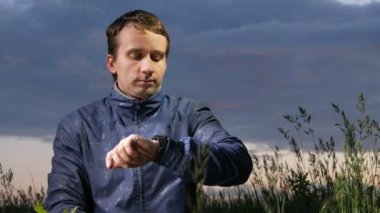 Sevimli genç bir adam bir smartwatch iletilerde sahada günbatımında denetler. Bir arka plan üzerinde güzel gökyüzü.