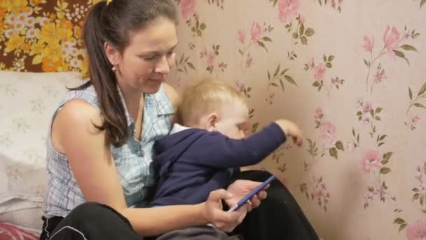 Молодая женщина с сыном сидит дома на диване с мобильным телефоном. Ребенок хочет выйти из объятий — стоковое видео