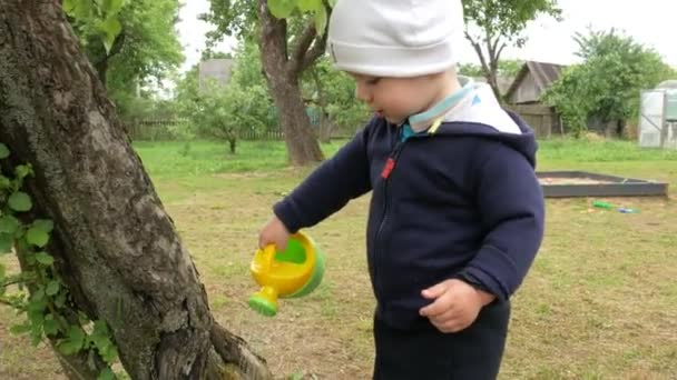 Um menino bonito derrama uma árvore grande de uma lata molhando pequena. Uma criança de bicicleta azul e um chapéu branco no jardim dos pais — Vídeo de Stock