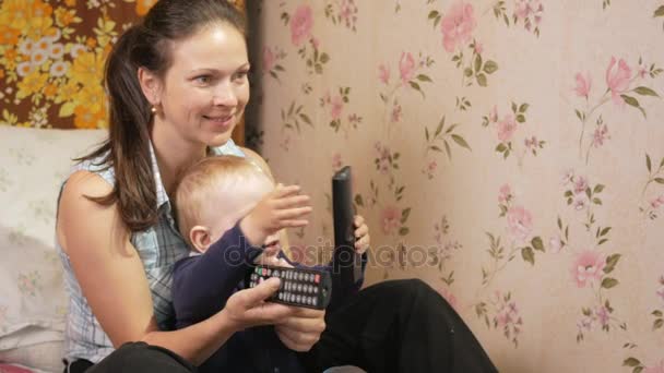年轻女子和她的儿子坐在家里在沙发上用遥控器从电视看屏幕用心 — 图库视频影像
