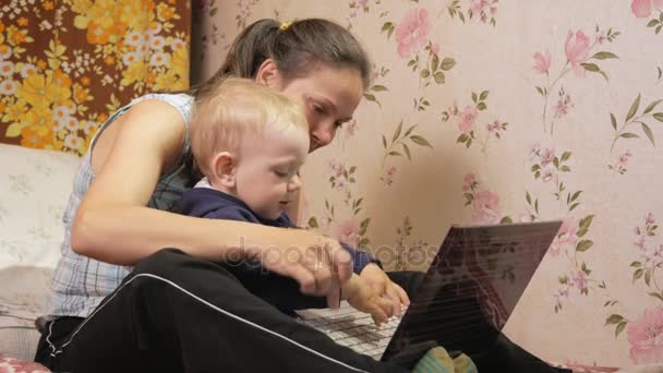 Giovane donna con suo figlio seduto a casa sul divano con un computer portatile. Guarda attentamente lo schermo e premi i tasti . — Video Stock