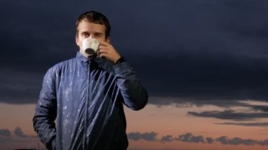 Sevimli genç biri sıcak bir içecek sahada günbatımında içer. Bir arka plan üzerinde güzel gökyüzü.