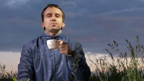 Un ragazzo carino beve un drink caldo al tramonto sul campo. Bel cielo su uno sfondo . — Video Stock