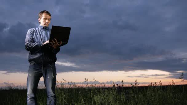 Netter junger Mann, der bei Sonnenuntergang auf dem Feld am Laptop arbeitet. schöner Himmel auf einem Hintergrund. — Stockvideo