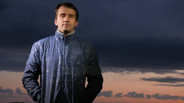 Un joven guapo mirando la cámara al atardecer en el campo. Hermoso cielo sobre un fondo . — Vídeos de Stock