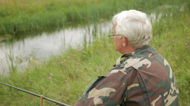 Un anciano está pescando en un pequeño río en el verano. Utiliza una caña de pescar y gusanos. Bosque y hierba verde alta — Vídeos de Stock