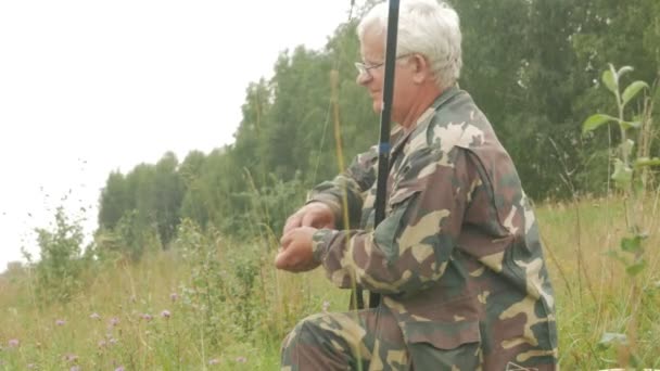 Um homem idoso está pescando em um pequeno rio no verão. Usa uma vara de pesca e vermes. Floresta e grama verde alta — Vídeo de Stock