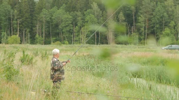 Um homem idoso está pescando em um pequeno rio no verão. Usa uma vara de pesca e vermes. Floresta e grama verde alta — Vídeo de Stock