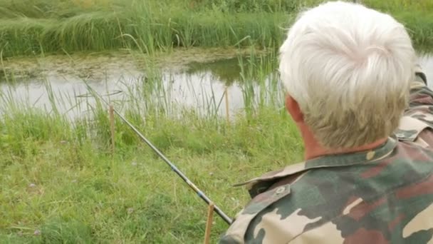 Un anciano está pescando en un pequeño río en el verano. Utiliza una caña de pescar y gusanos. Bosque y hierba verde alta — Vídeos de Stock