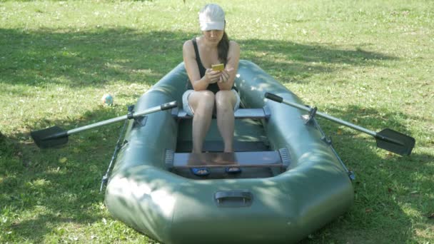 Una mujer bonita se sienta en un bote inflable cerca de la casa y marca el teléfono. Pantalones cortos y una gorra blanca de béisbol en la cabeza — Vídeos de Stock