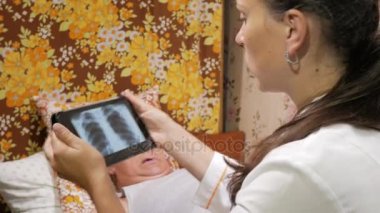 Kadın doktor hasta evde ziyaret etti. Bir tablet bilgisayarda röntgen sonuçlarını gösterir. Adam kanepede yatıyor.