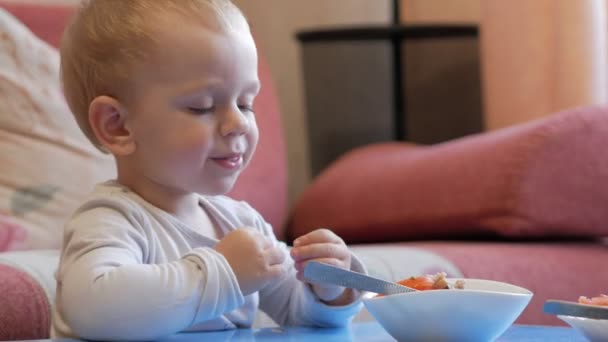 Um menino bonito come mingau de cereal com pedaços de carne em uma mesa de crianças. mobiliário doméstico — Vídeo de Stock