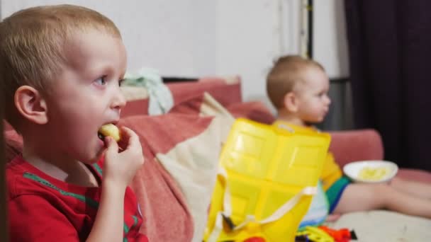 Die Brüder sitzen zu Hause auf dem Sofa und schauen fern. Maisstangen essen und lächeln — Stockvideo