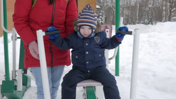 A cute little boy and a young mother are engaged in outdoor exercise equipment. Its hard for a kid to reach the handles of equipment — Stock Video