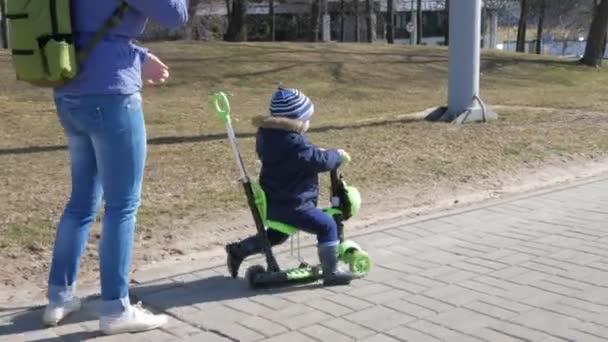 Une jeune mère et un garçon sortent. Le gamin skate assis sur le scooter — Video