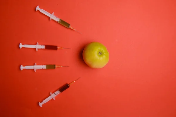 Chemical additives in food or genetically modified fruit concept. Green apple with syringes of chemicals. Isolated on red background.