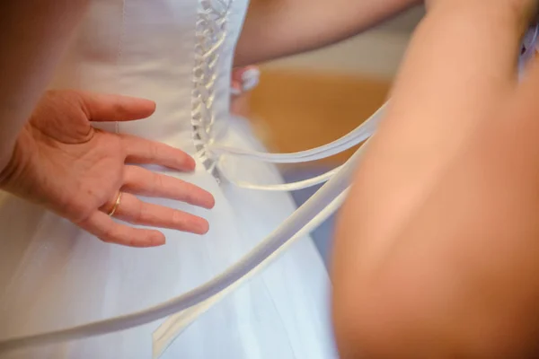 Dama de honra ajudando noiva esbelta amarrando seu vestido branco do casamento, abotoando no padrão de renda delicada com saia macia na cintura. Manhã nupcial preparação detalhes recém-casados. Momentos do dia do casamento, desgaste. — Fotografia de Stock