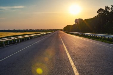 Akşam otoyolda. Kırsal kesimde asfalt yol, ekili tarlalar ve yandan orman var..