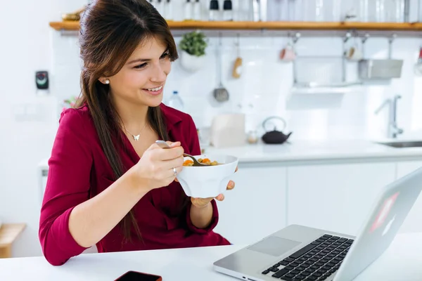 Mulher bonita trabalhando com laptop enquanto come na cozinha . — Fotografia de Stock