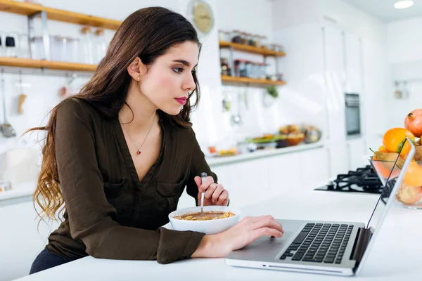 Hermosa mujer joven que trabaja con el ordenador portátil mientras come en la cocina . — Foto de Stock