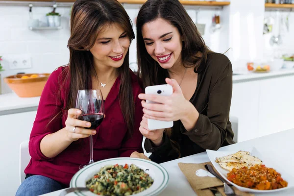 Two young women using mobile phone while eating in the kitchen. — Stock Photo, Image