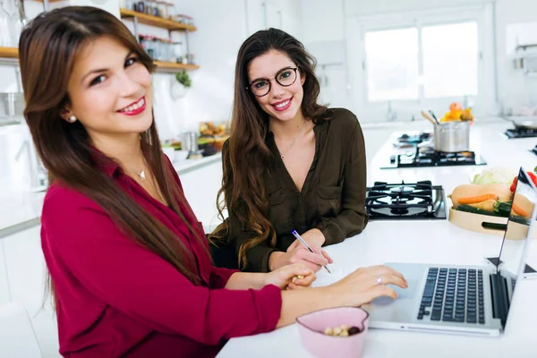 Deux belles jeunes femmes travaillant avec un ordinateur portable dans la cuisine . — Photo