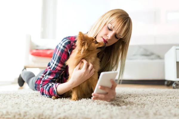 Belle jeune femme avec son chien utilisant un téléphone portable à la maison . — Photo