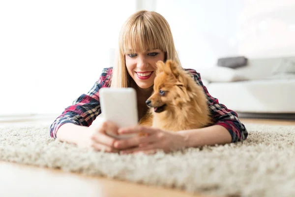 Belle jeune femme avec son chien utilisant un téléphone portable à la maison . — Photo