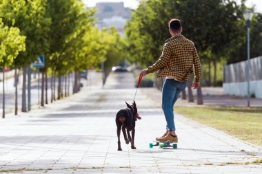 Yakışıklı genç adam köpeğiyle parkta kaykay.