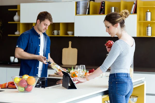 Bonito jovem abrindo garrafa de vinho, enquanto sua esposa usando tablet digital na cozinha . — Fotografia de Stock