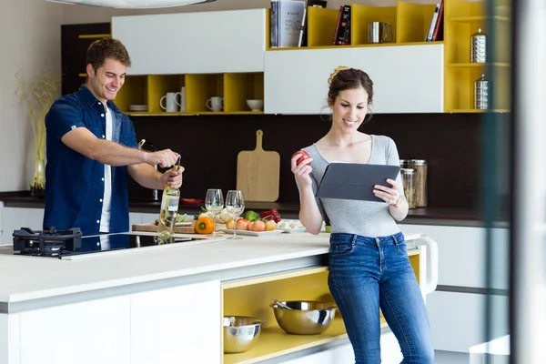 Schöner junger Mann öffnet Weinflasche, während seine Frau digitales Tablet in der Küche benutzt. — Stockfoto