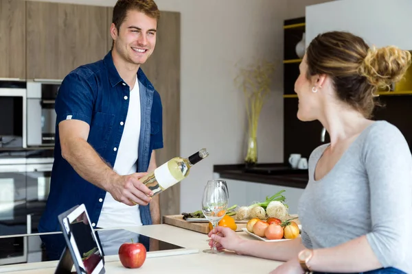 Bonito jovem abrindo garrafa de vinho, enquanto sua esposa usando tablet digital na cozinha . — Fotografia de Stock