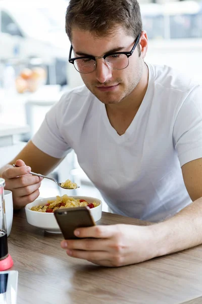 Csinos, fiatal férfi, miközben élvezi a reggelit, a konyhában a mobiltelefon használata. — Stock Fotó