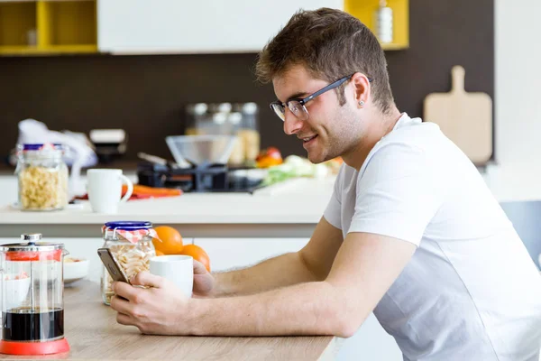 Bonito jovem usando seu telefone celular enquanto desfruta do café da manhã na cozinha . — Fotografia de Stock