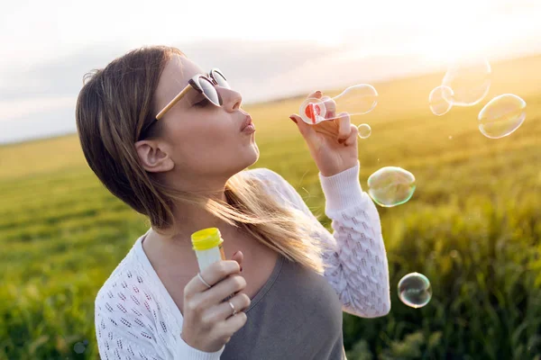 Schöne junge Frau pustet Seifenblasen auf einem Feld. — Stockfoto