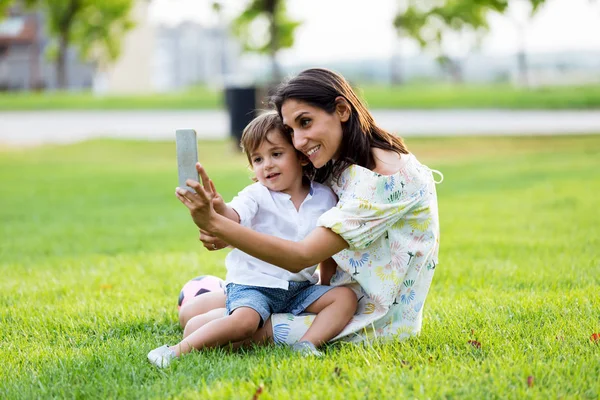 Bela jovem mãe com seu filho tirando uma selfie no parque . — Fotografia de Stock