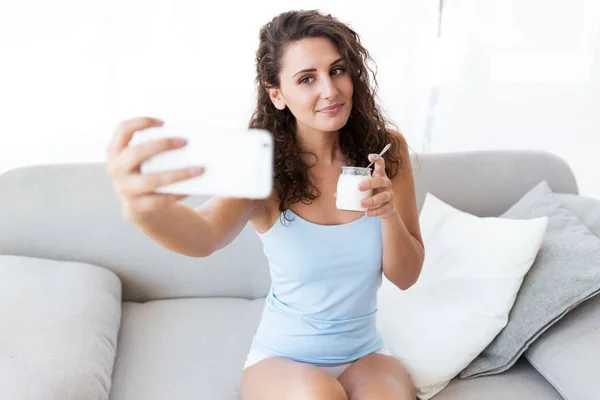 Mulher bonita tomando uma selfie enquanto come iogurte em casa . — Fotografia de Stock