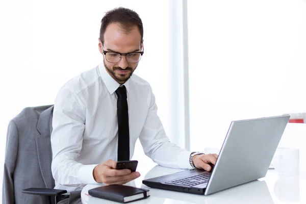 Komea nuori mies työskentelee kannettavan tietokoneen ja matkapuhelimen kanssa toimistossa . — kuvapankkivalokuva