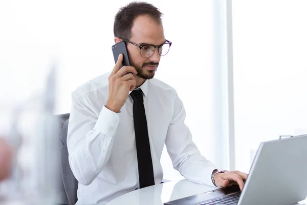 Joven guapo que trabaja con el ordenador portátil y el teléfono móvil en la oficina . — Foto de Stock