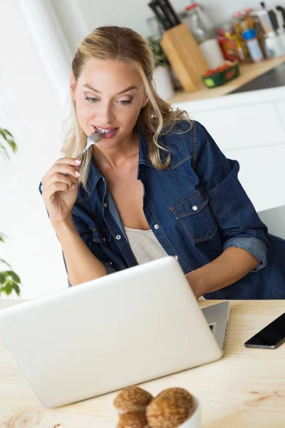 Beautiful young woman using her laptop and eating yogurt at home — Stock Photo, Image