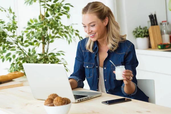 Mulher bonita usando seu laptop e comer iogurte em casa — Fotografia de Stock
