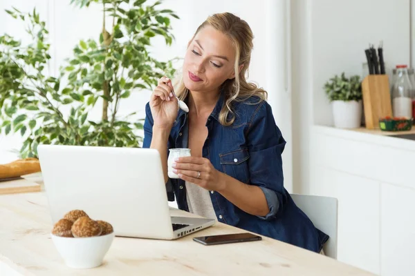 Beautiful young woman using her laptop and eating yogurt at home — Stock Photo, Image