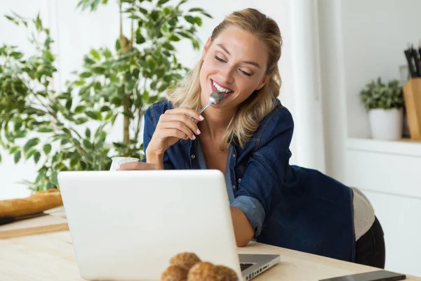 Mulher bonita usando seu laptop e comer iogurte em casa — Fotografia de Stock