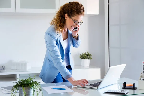 Młoda kobieta biznesu, rozmowy na telefon komórkowy, używając swojego laptopa w biurze. — Zdjęcie stockowe