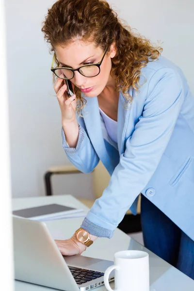 Mujer joven de negocios hablando en el teléfono móvil mientras usa su computadora portátil en la oficina . — Foto de Stock