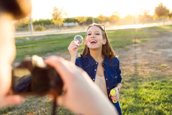 Handsome young man taking photo of his girlfriend making soap bubbles in the park. — Stock Photo, Image